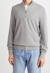 Man i en ljusgrå halvzip-tröja över en vit skjorta, tillsammans med ljusblå denimjeans, stående framför en enfärgad bakgrund.