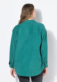 Chemise en tissu vert doux avec une coupe décontractée, des manches longues et des poignets boutonnés. Comprend un empiècement au dos et un ourlet arrondi pour un look décontracté.