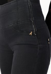 Crne traperice s visokim strukom, zatvaranjem na zip i zlatnim akcentom u obliku pčele na džepu. Tkanina ima elegantnu, glatku teksturu.