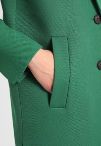 Grüne strukturierte Jacke mit einer Hand einer Person, die in die Tasche gesteckt ist, und die den Stoff und die Designdetails des Kleidungsstücks zeigt.