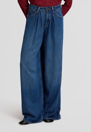Person trägt weite blaue Denim-Jeans kombiniert mit schwarzen Schuhen und einem roten langärmligen Oberteil, steht mit Händen in den Hosentaschen.