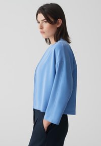 Veste courte bleu clair en tissu lisse, avec un col rond, des épaules tombantes et des manches longues. Design minimal sans fermetures.