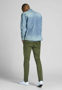 Giacca di jeans blu chiaro con un aspetto sbiadito, abbinata a pantaloni slim-fit verde oliva e sneaker bianche, vista da dietro.