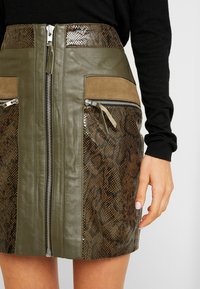 Jupe en cuir olive avec des accents en imprimé serpent, dotée d'une fermeture éclair centrale, de deux poches zippées sur les côtés et d'une finition texturée.