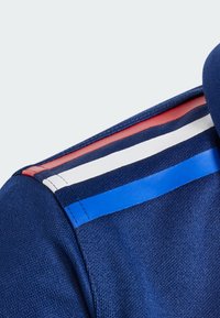 Námořní sportovní košile s límcem, který má texturovanou tkaninu a barevný pruhový design v červené, bílé a modré barvě napříč ramenem.