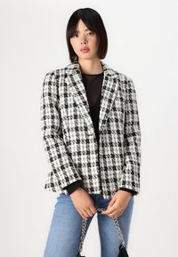 Black and white checked blazer in textured fabric, featuring a single-button closure, notched lapels, and a loose fit. Paired with denim.