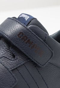 Sneaker in pelle blu scuro con finitura tessurizzata, dotato di una strap in velcro, dettagli cuciti e un'etichetta logo visibile nella parte superiore.