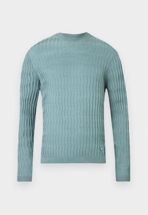 Gaiši zils džemperis no mīkstas auduma ar reljefu viļņu rakstu, garām piedurknēm un apaļu kaklu. Nav ievērojamu aparatūras detaļu vai akcentu.