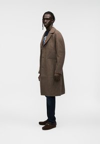 Καφέ μάλλινο παλτό με γιακά με εγκοπές, ένα κουμπί και δύο πλαϊνές τσέπες. Συνδυασμένο με σκούρο τζιν και καφέ παπούτσια τύπου slip-on.