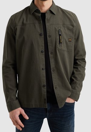 Homme portant une chemise verte à manches longues déboutonnée par-dessus un t-shirt noir et un jean bleu, avec une main dans la poche et l'autre détendue.