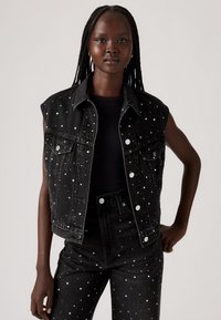 Zwarte denim mouwloze jas met zilveren en gouden studs, gecombineerd met bijpassende zwart gestoffeerde jeans, tegen een neutrale achtergrond.