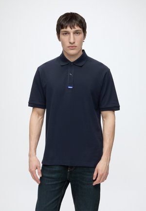 Junger Mann trägt ein marineblaues kurzärmeliges Poloshirt mit kleinem blauem Hugo-Logo auf der Brust, steht vor einem schlichten hellen Hintergrund.