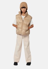 Enfant portant un bonnet en tricot beige, un gilet matelassé brillant beige, une chemise à manches courtes blanche, un pantalon large beige et des chaussures noires, se tenant avec les mains dans les poches.