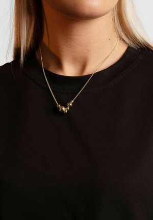 Gold necklace with round beads on a fine chain, worn over a black t-shirt. Necklace is layered, with varying bead sizes and a minimalist design.
