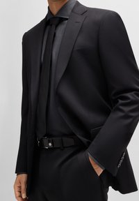 Schwarzer maßgeschneiderter Anzug mit satinierter Oberfläche, kombiniert mit einer passenden Krawatte und einem strukturierten Hemd. Das Outfit wird durch einen schwarzen Gürtel mit silbernen Verschlüssen abgerundet.