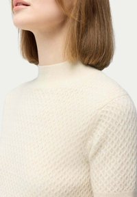 Stickad krämfärgad pullover med en texturerad diamantmönster, med hög krage och korta ärmar, tillverkad av mjukt material.