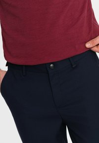 Pantaloni blu navy realizzati in tessuto texturizzato, con chiusura a bottone metallico e tasche frontali, abbinati a una camicia leggera bordeaux.