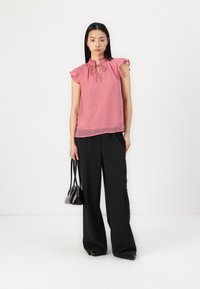 Top rosa con pois, maniche arricciate e scollatura a occhiello, abbinato a pantaloni neri a vita alta e gamba larga, insieme a una piccola borsa nera.