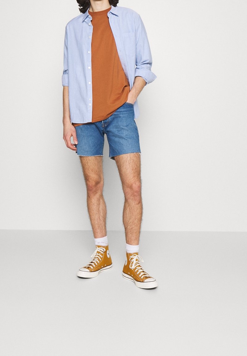 Personne portant un t-shirt orange, une chemise bleu clair ouverte avec boutons, un short en jean, des chaussettes blanches et des baskets montantes jaune moutarde, debout avec une main dans la poche.