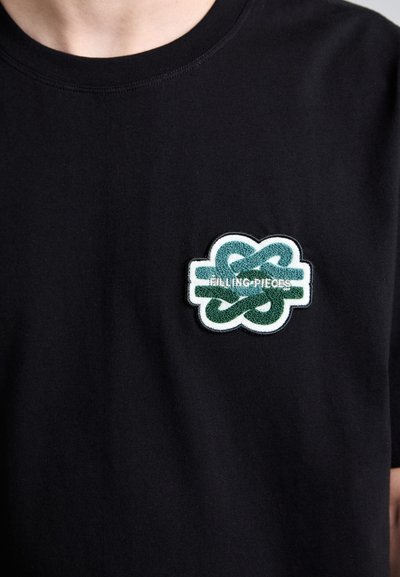 Black cotton t-shirt featuring a green and white embroidered logo patch on the left chest area. Rounded neckline and short sleeves.