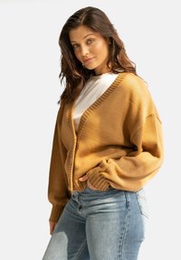 Musztardowy kardigan z dekoltem w serek, prążkowanymi brzegami i oversize'owymi rękawami, noszony na białej koszulce i zestawiony z dopasowanymi niebieskimi dżinsami.