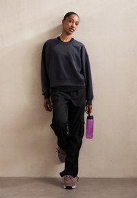 Fekete, oversized kapucnis póló, fekete cargo nadrág zsebekkel, lila vízpalack és világos rózsaszín sportcipő. A modell egy semleges színű falnak dől.