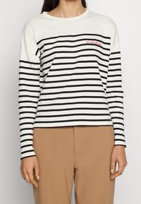 Långärmad randig topp i vitt och svart med en figurnära design. Har en röd broderad "måste ha" text på bröstet. Mjuk tyg.