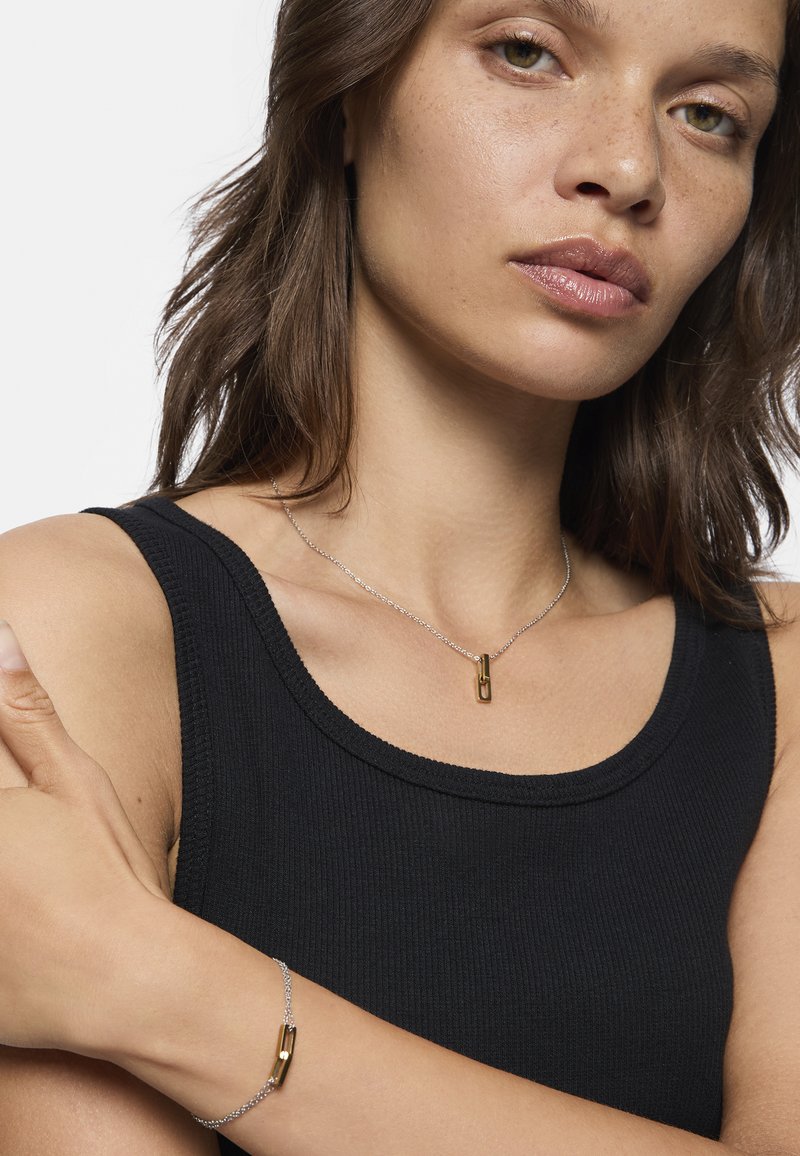 Silberne Halskette mit einem Anhänger aus goldfarbenem Clip, dazu passende Armbänder mit goldenen Akzenten, getragen über einem schwarzen, gerippten Tank-Top. Glatte Textur, minimalistisches Design.