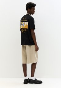 Fekete grafikus póló hátoldalán absztrakt formákkal. Bézs színű, térd fölé érő sorttal és fekete loaferekrel párosítva, amelyekhez fehér zoknik tartoznak.