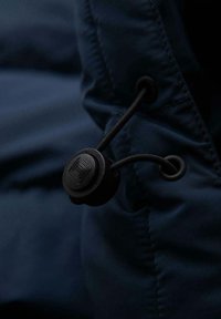 Il tessuto blu navy presenta un primo piano di un bloccaggio a toggle nero e corda, con una texture liscia e un design minimalista per la regolazione.