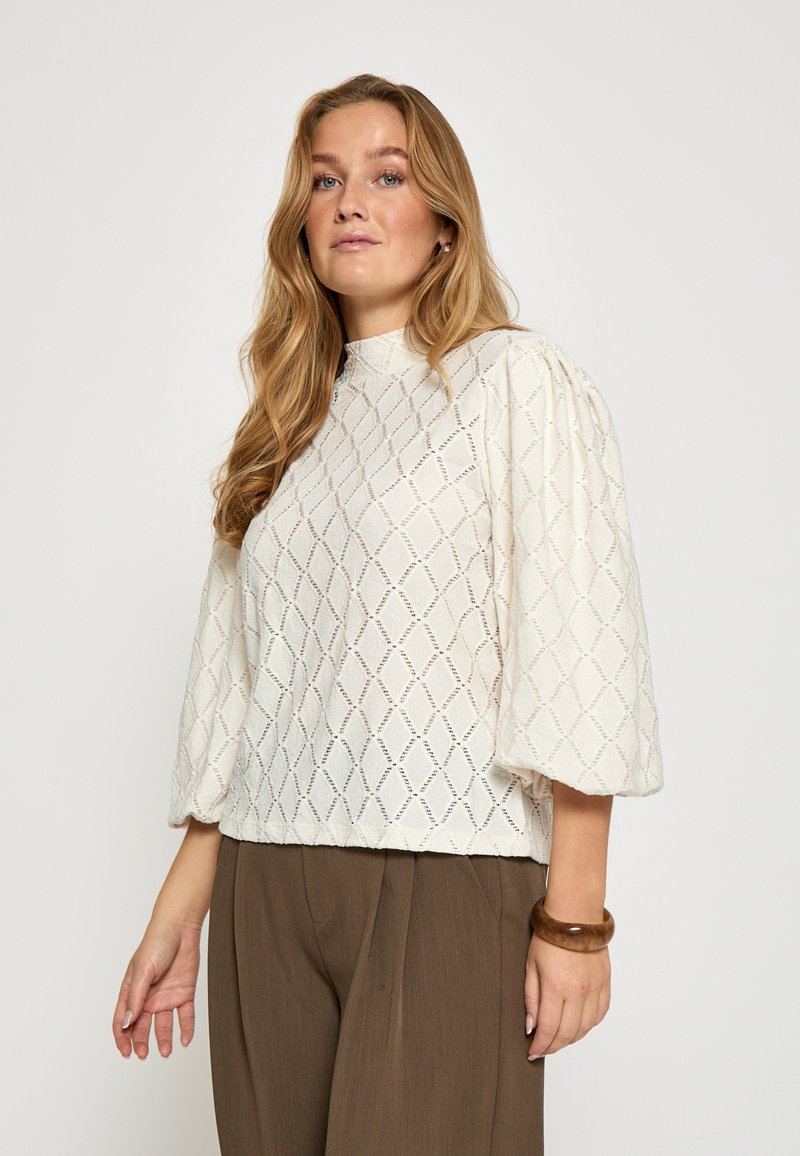 Biely top s nadýchanými rukávmi s textúrovaným diamantovým vzorom, vysokým golierom a pohodlným strihom, v kombinácii s hnedými širokými nohavicami.
