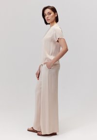 Jasnobeżowa jedwabna bluzka z krótkimi rękawami, zestawiona z spodniami o szerokich nogawkach. Spodnie mają boczne kieszenie. Modelka stoi w profilu, nosząc płaskie sandały.