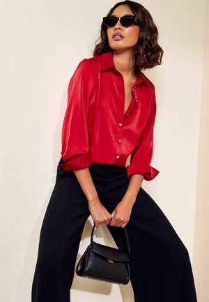 Vrouw met zwarte zonnebril, rode satijnen blouse, zwarte wijde broek, die een kleine zwarte handtas vasthoudt tegen een effen lichte achtergrond.