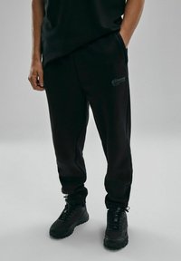 Czarne długie spodnie dresowe w dopasowanym kroju, wykonane z miękkiego materiału, z bocznymi kieszeniami, subtelnym detalem logo i w zestawie z czarnymi, teksturowanymi sneakersami.