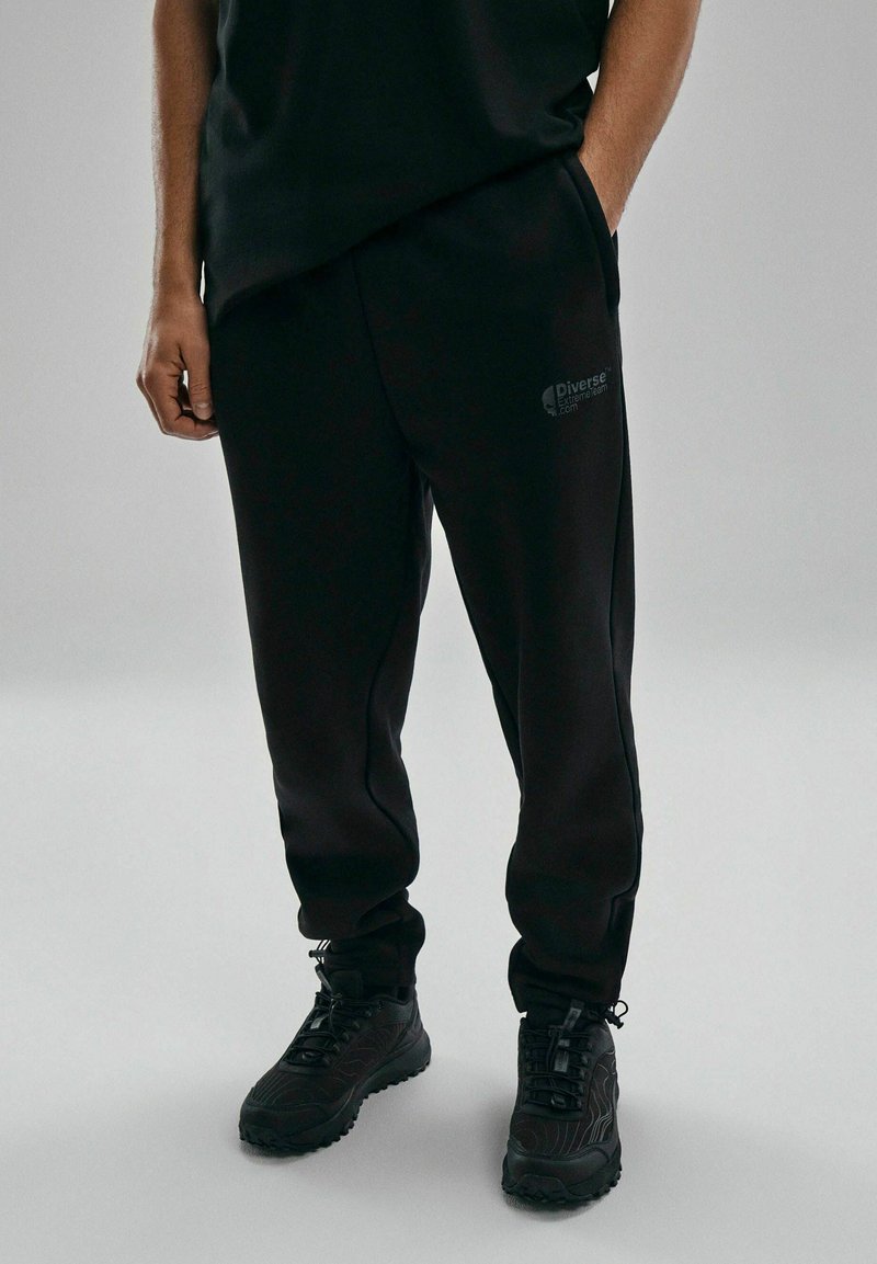 Czarne długie spodnie dresowe w dopasowanym kroju, wykonane z miękkiego materiału, z bocznymi kieszeniami, subtelnym detalem logo i w zestawie z czarnymi, teksturowanymi sneakersami.