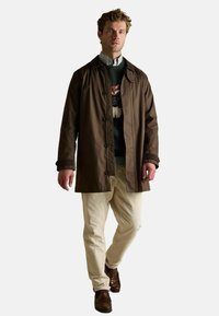 Brączowy wodoodporny płaszcz z zapięciem na guziki, noszony na wzorzystym swetrze i koszuli z kołnierzykiem, w połączeniu z kremowymi spodniami z kordurowego materiału i brązowymi skórzanymi butami.