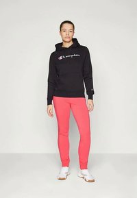 Sieviete stāv tērpusies melnā Champion kapucē, spilgti rozā legingos un baltās čībās uz vienkārša gaiša fona.