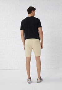 Sort t-shirt med korte ærmer, beige shorts med baglomme og grå/sølv sportssko, set fra baggrunden mod en hvid murstensvæg.
