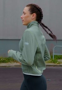 Mulher jovem com cabelo trançado a correr ao ar livre, vestindo uma jaqueta texturada verde e leggings pretas em um ambiente urbano.