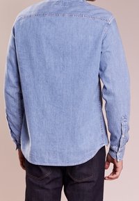 Ljusblå denimskjorta med långa ärmar, krage med knappar och rundad nederkant. Har sömdetaljer och knappar vid ärmslut.