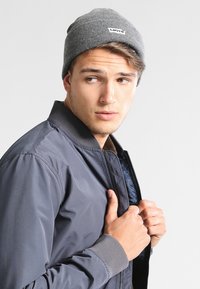 Gorro de punto gris con una etiqueta de logo, combinado con una chaqueta bomber azul oscuro con cierre de cremallera y puños acanalados, llevada de forma casual.