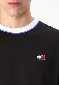 Juodas trumpas rankovėmis marškinėliai su apvaliu kaklu, papuošti balta ir mėlyna apdaila. Išsiuvinėtas logotipas raudona, balta ir mėlyna spalvomis ant krūtinės.