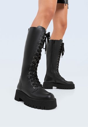 Kobieta w wysokich do kolan czarnych skórzanych botkach na platformie z grubymi podeszwami na jasnym tle.
