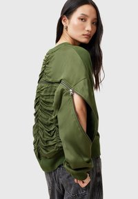 Olivgrön sweatshirt med rynkad baksida, oversize passform och dragkedjor på ärmarna. Har mjukt, slätt material och kontrasterande texturer.