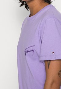 T-shirt di cotone lilla con maniche corte, caratterizzata da un logo ricamato "Tommy" in viola scuro sul petto e un dettaglio di marca sulla manica.