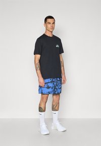T-shirt in cotone nero con piccolo logo turchese, abbinato a pantaloni corti neri e blu a fantasia. Calzini alla caviglia bianchi e sneakers bianche completano il look.