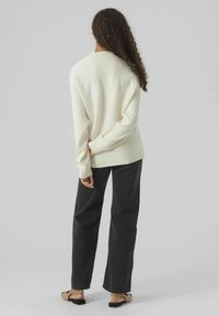 Cremfarbener, gerippter Strickpullover mit breitem Kragen, kombiniert mit lässig geschnittenen schwarzen Jeans und flachen Sandalen, von hinten gesehen.