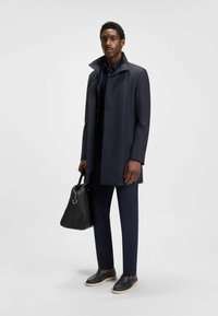 Cappotto impermeabile blu navy con una leggera lucentezza, colletto abbottonato, abbinato a pantaloni coordinati e scarpe slip-on, con una borsa in pelle nera.