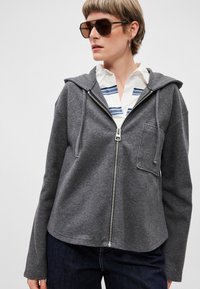 Sivá mikina na zips vyrobená z mäkkej tkaniny, s prednou kapsou, kapucňou a dlhými rukávmi, prevlečená cez biele tričko s modrými pruhmi.