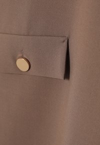 Brown fabric with a smooth texture featuring a patch pocket secured by a gold button detail. Focus on tailored design and color consistency.